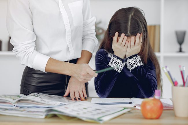a little girl hiding her face with her hands, an older woman asking her to write with a pencil